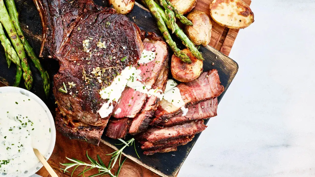 En saftig tomahawk steak skuren i skivor, serverad med rostade potatisar, sparris och en kr&auml;mig s&aring;s.