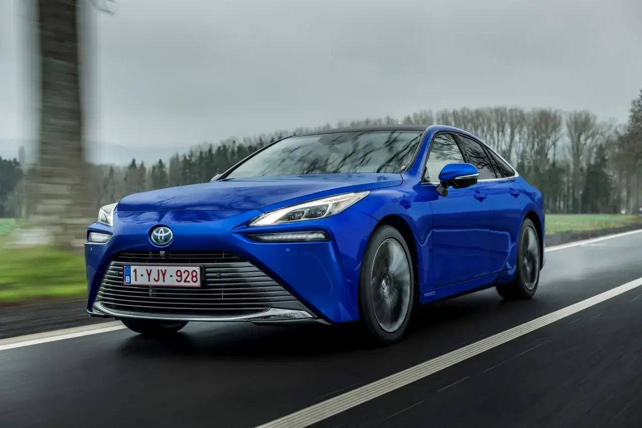 Niebieska Toyota Mirai jedzie po mokrej drodze, otoczona drzewami.