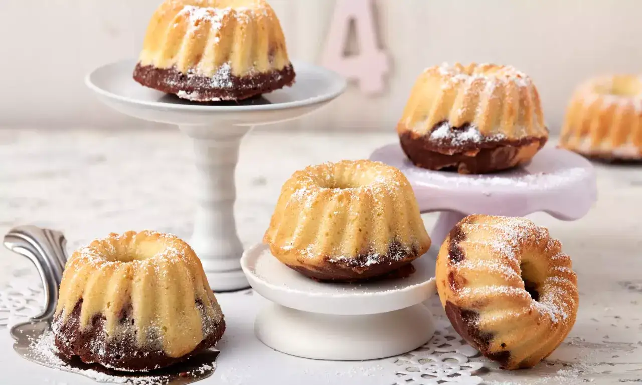 Mehrere kleine, mit Puderzucker best&auml;ubte Mini Gugelhupf-Kuchen mit Schokoladenboden stehen auf verschiedenen Etageren.