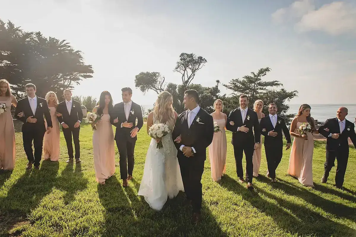 A wedding party walks in the sun. The bride and groom are in the center, surrounded by bridesmaids in blush dresses and groomsmen in black tie.