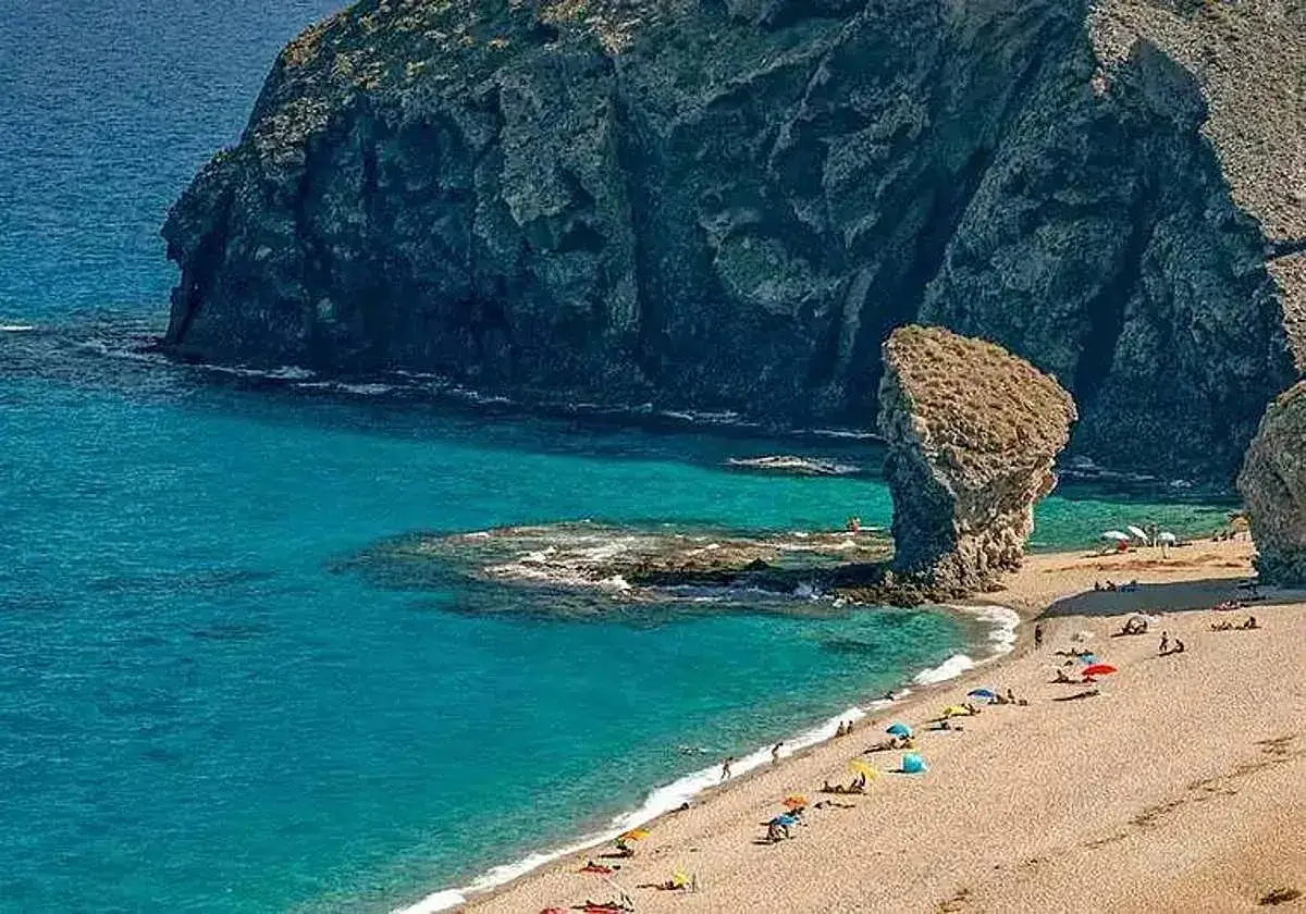 Aguas turquesas bañan las calas en Vera, con formaciones rocosas imponentes y gente disfrutando del sol bajo sombrillas.