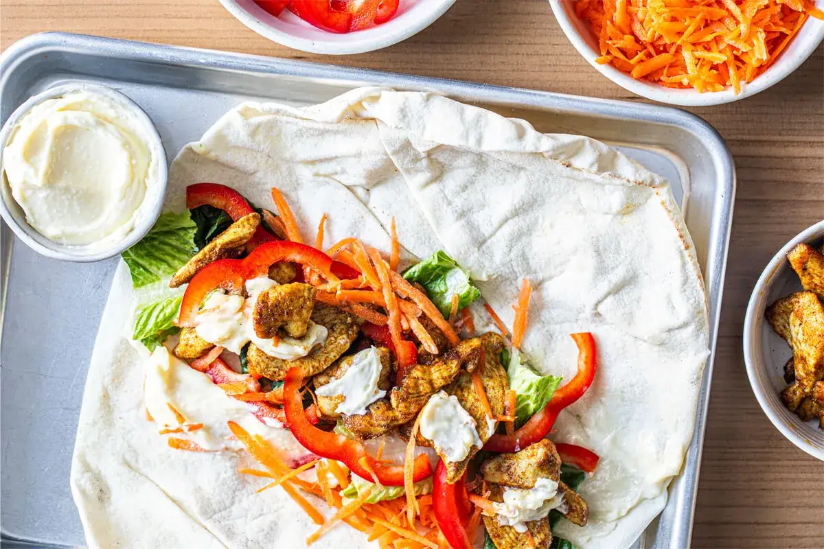 En kyckling shawarma med grönsaker och sås på ett tunnbröd, redo att rullas. Bredvid finns en skål med vit sås och en skål med strimlade morötter.