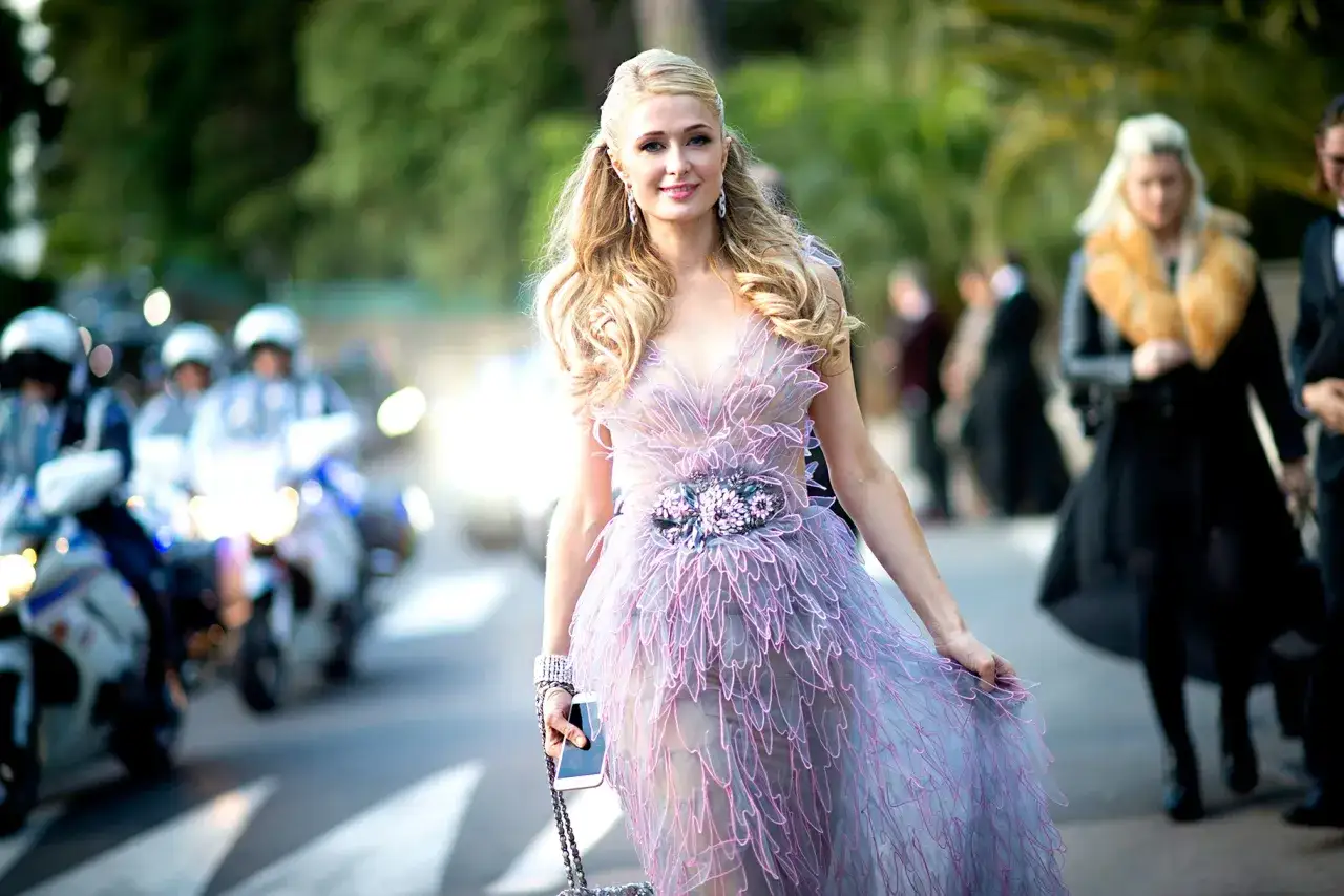 Paris Hilton w zwiewnej sukni, jak z filmu, idzie ulicą, otoczona przez policję na motocyklach.