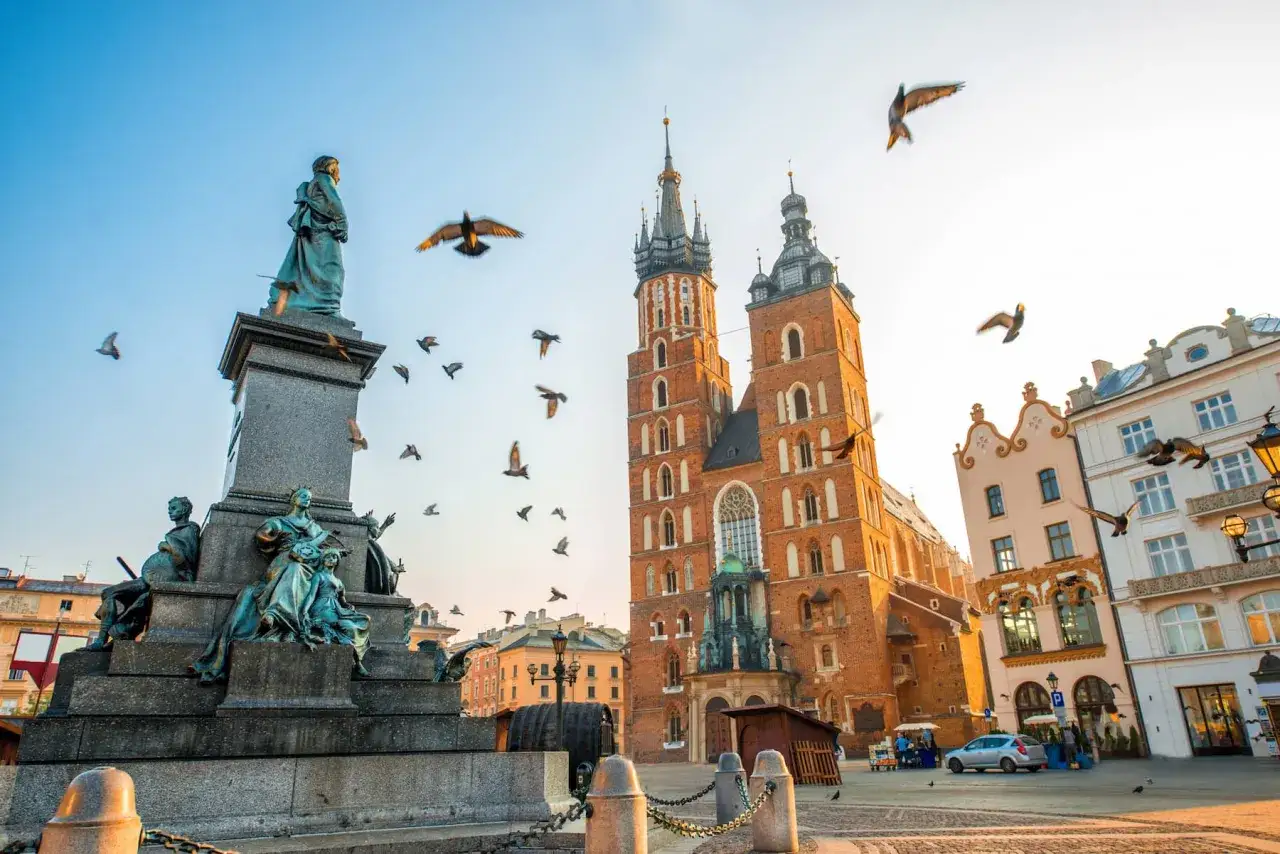 A flock of pigeons flies around the monument and St. Mary's Basilica in Krakow's Main Market Square, a quintessential Krakow travel scene.