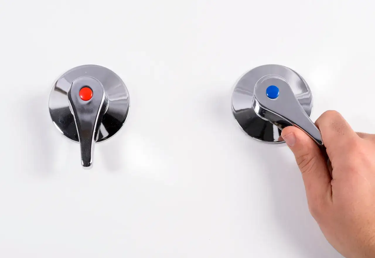 Dos grifos cromados, uno con punto rojo y otro con punto azul, sobre fondo blanco. Una mano ajusta la llave de paso de agua general del grifo azul.