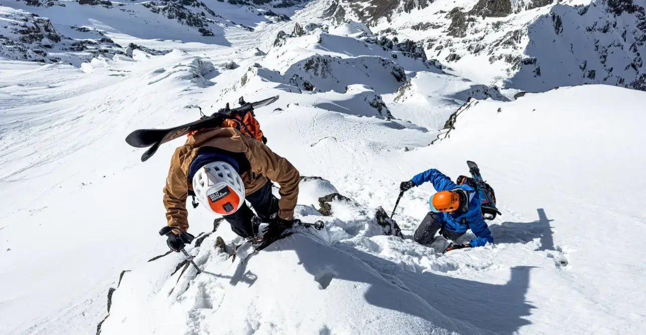 Zwei Skibergsteiger k&auml;mpfen sich durch steiles, schneebedecktes Gel&auml;nde. Einer tr&auml;gt ein Snowboard auf dem R&uuml;cken, der andere einen Skistock. Sie nutzen ihre H&auml;nde und St&ouml;cke, um die rupal wand zu erklimmen.