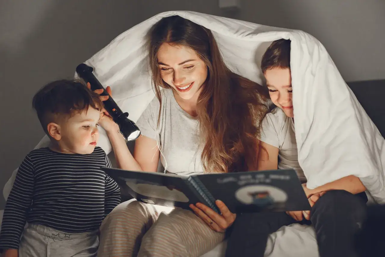 Madre lee un cuento a sus hijos bajo una manta con linterna. Perfectas frases del D&iacute;a del Libro para ni&ntilde;os.