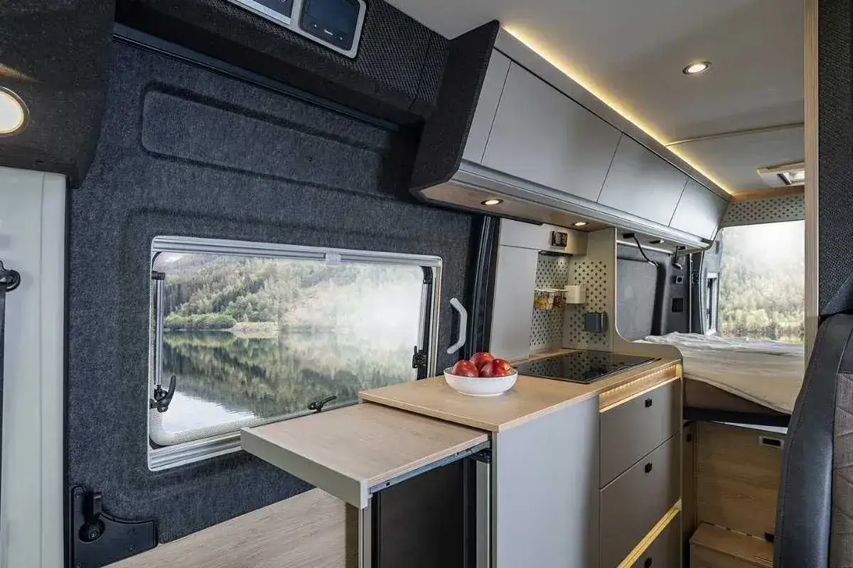 Cocina de un camper garage con vista a un lago. Muebles modernos, encimera con vitrocerámica y cama visible.