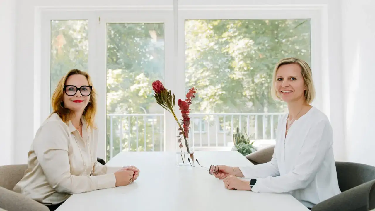 Zwei Frauen am Tisch, diskutieren über Ehevertrag ja oder nein. Blumen in Vase, Fenster mit Bäumen im Hintergrund.