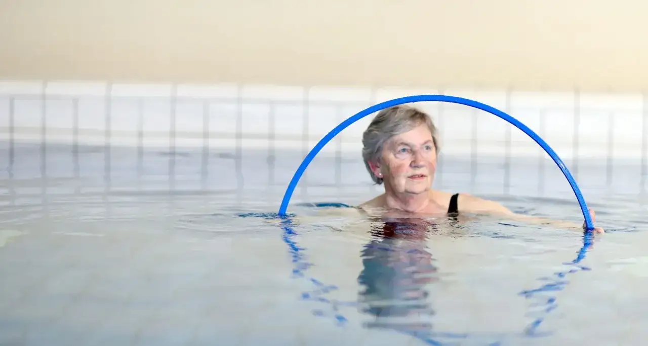 &Auml;ltere Frau mit grauem Haar in einem Schwimmbecken, die mit einem blauen Ring &uuml;bt. Balneotherapie bei Arthrose hilft ihr, sich zu bewegen.