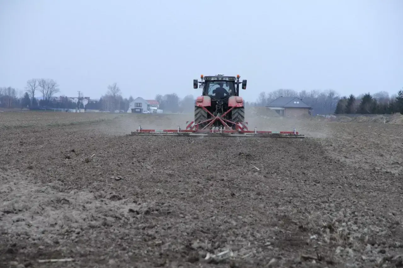 Czerwony traktor z agregatem uprawowym pracuje na polu. To przygotowanie ziemi pod przyszłe plony, być może na dzierżawę.