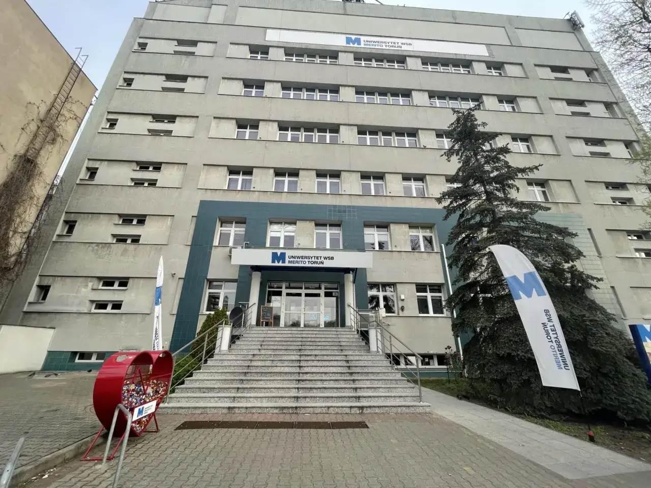 Budynek Uniwersytetu WSB Merito Toruń z charakterystycznym wejściem i schodami. Obok serce-śmietnik i flagi z logo uczelni.