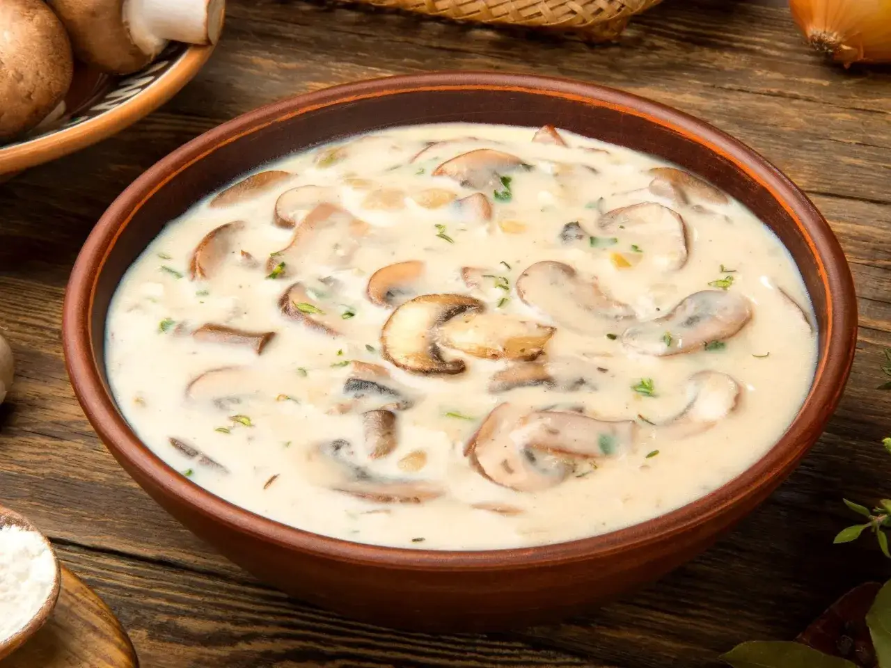 Aksamitna zupa pieczarkowa w glinianej misce, pełna kawałków grzybów i ziół. Obok leżą świeże pieczarki i cebula.