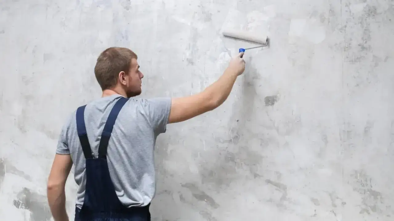 Mężczyzna w ogrodniczkach maluje wałkiem ścianę, przygotowując ją pod farbę. Dowiadujemy się, czym gruntować ściany, by uzyskać idealne wykończenie.