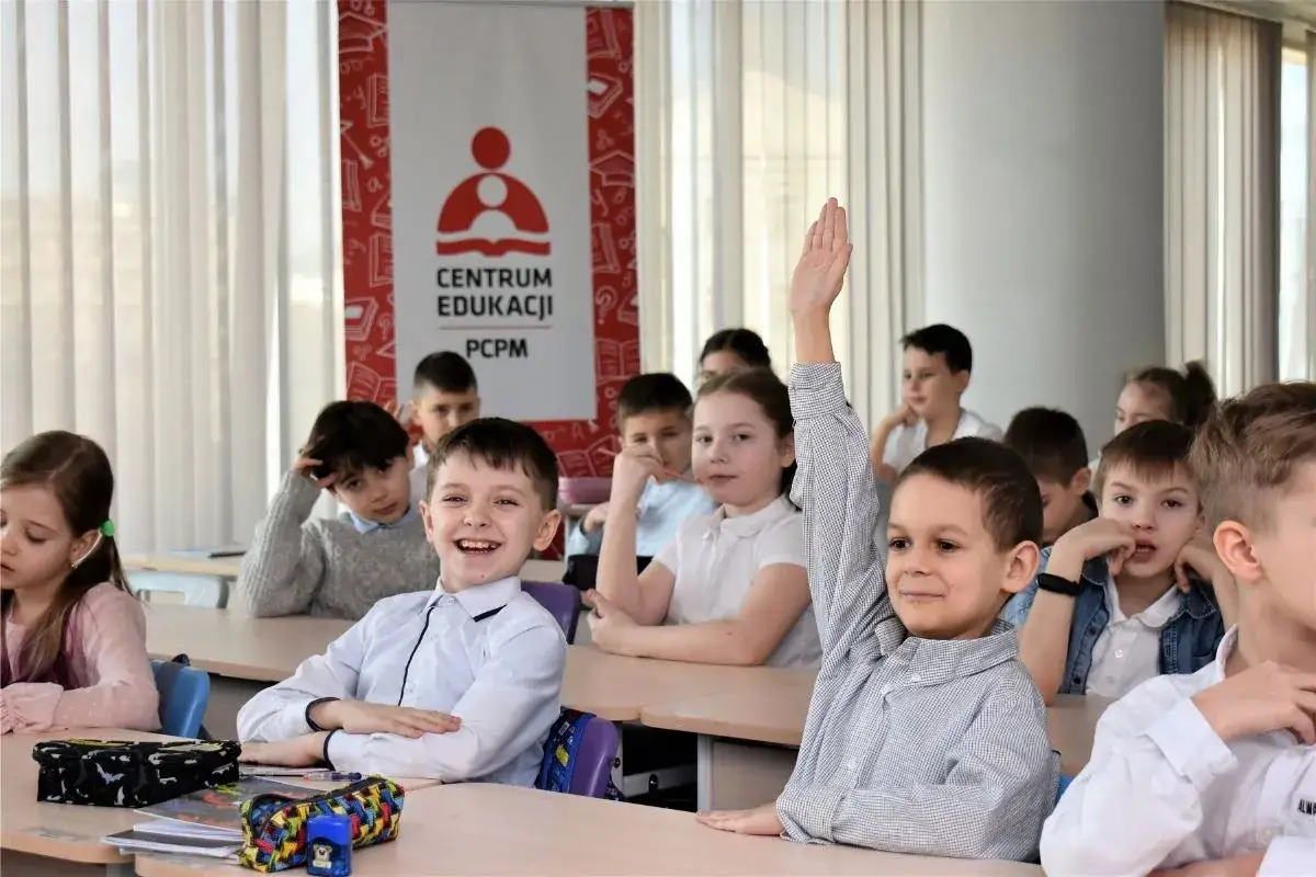 Uśmiechnięty chłopiec z podniesioną ręką, gotowy do odpowiedzi. Wszyscy uczniowie w klasie wypełniają swój obowiązek szkolny w Polsce.