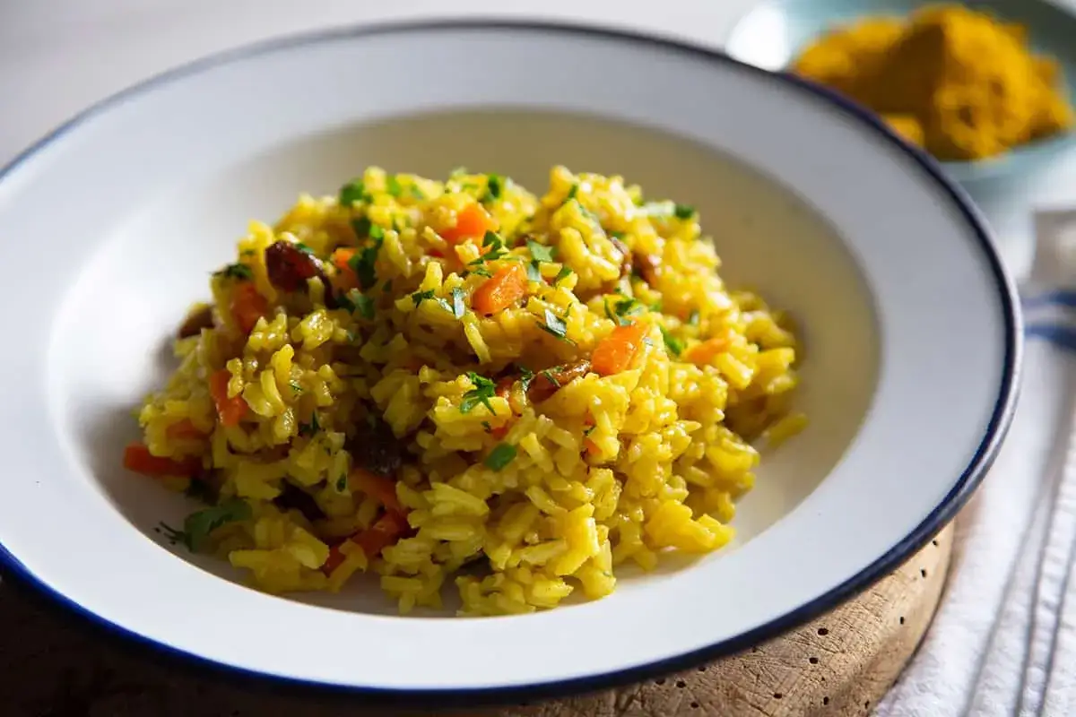 Un plato de arroz al curry con zanahoria y pasas, adornado con perejil fresco. Al fondo, un mont&iacute;culo de c&uacute;rcuma en polvo.