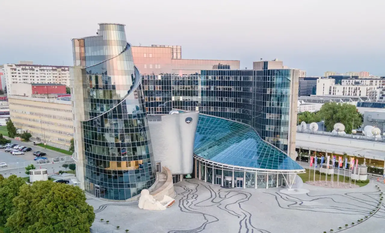 Nowoczesny budynek z przeszkloną fasadą, przypominający falę. W tle widać inne budynki, a na placu przed wejściem rzeźbę. To siedziba TVP w likwidacji.