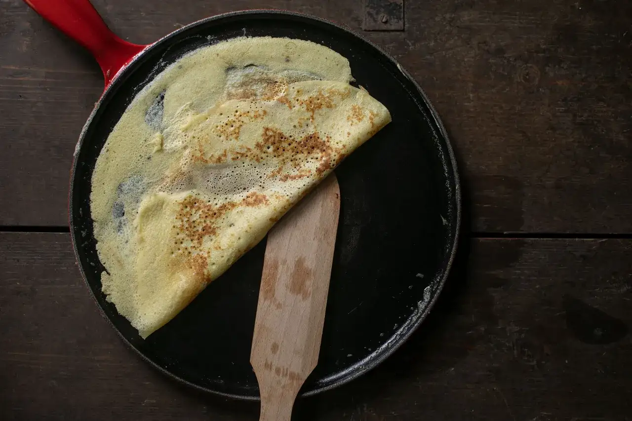 Ein goldbrauner Cr&ecirc;pe wird in einer Pfanne mit rotem Griff mit einem Holzspatel gewendet. Das perfekte Cr&ecirc;pes Rezept gelingt so.