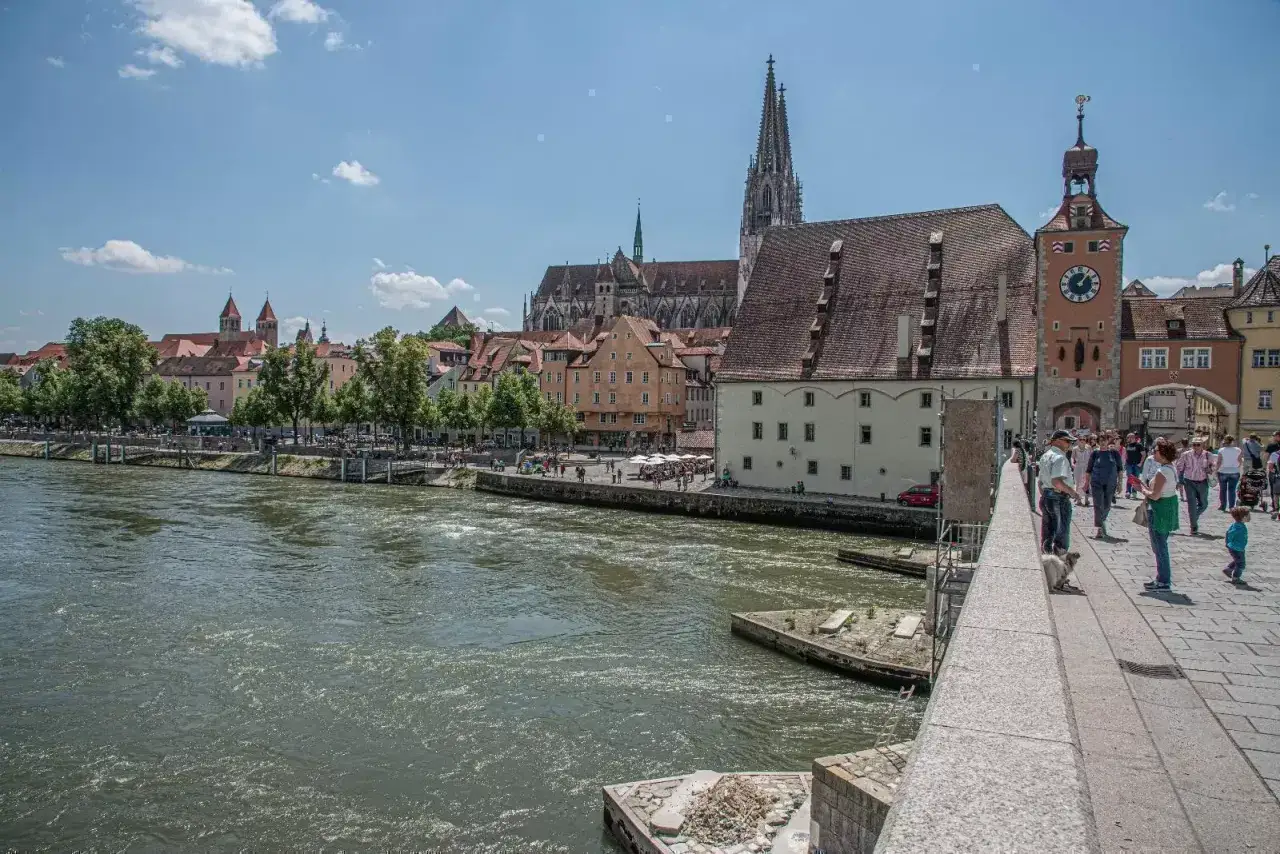 Regensburg sehenswertes: Die Donau fließt an der historischen Altstadt vorbei, mit dem Dom St. Peter und dem Steinerernen Turm. Menschen spazieren auf der Steinernen Brücke.