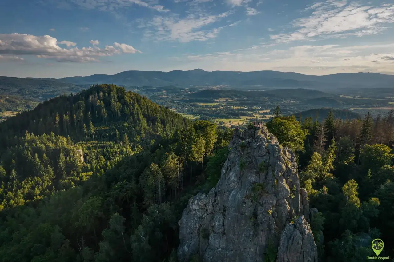 Widok na skaliste rudawy janowickie szczyty porośnięte lasem, z ludźmi na szczycie. W tle rozległa panorama gór i dolin.