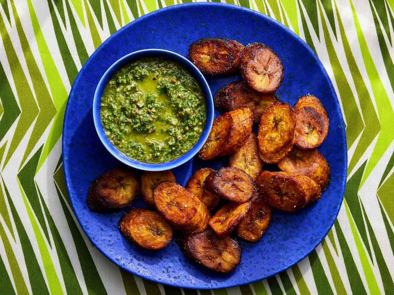 Plátano frito dorado y crujiente servido en un plato azul con un tazón de salsa verde vibrante.