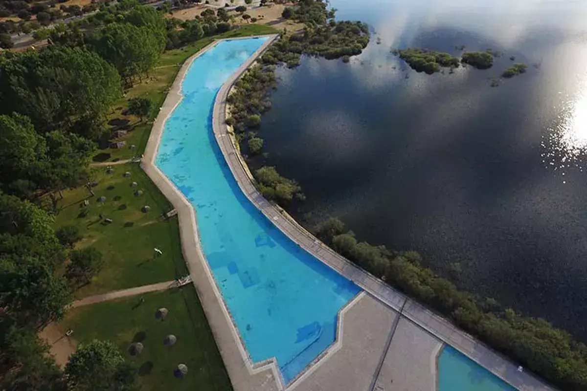 Vista aérea de la piscina natural de Riosequillo, con su agua turquesa bordeando un lago oscuro y rodeada de césped y árboles.