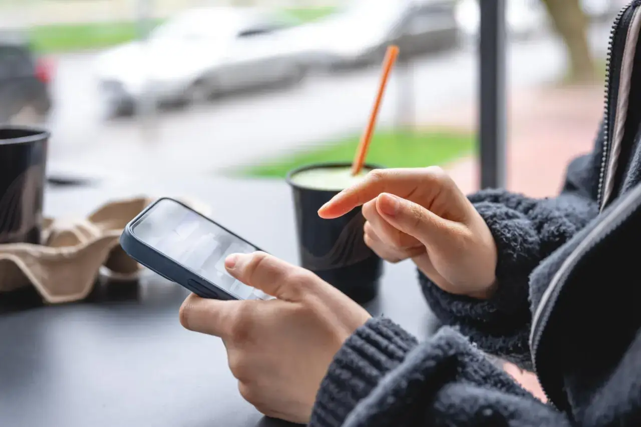Osoba korzysta z telefonu, sprawdzając, co to jest lokata mobilna. Obok napój i kubek.