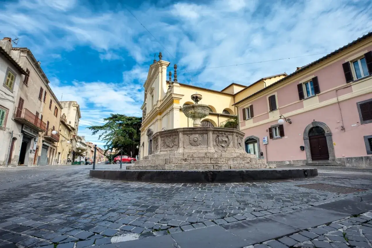 Fontana in pietra nel centro storico canino, con edifici colorati e cielo azzurro.