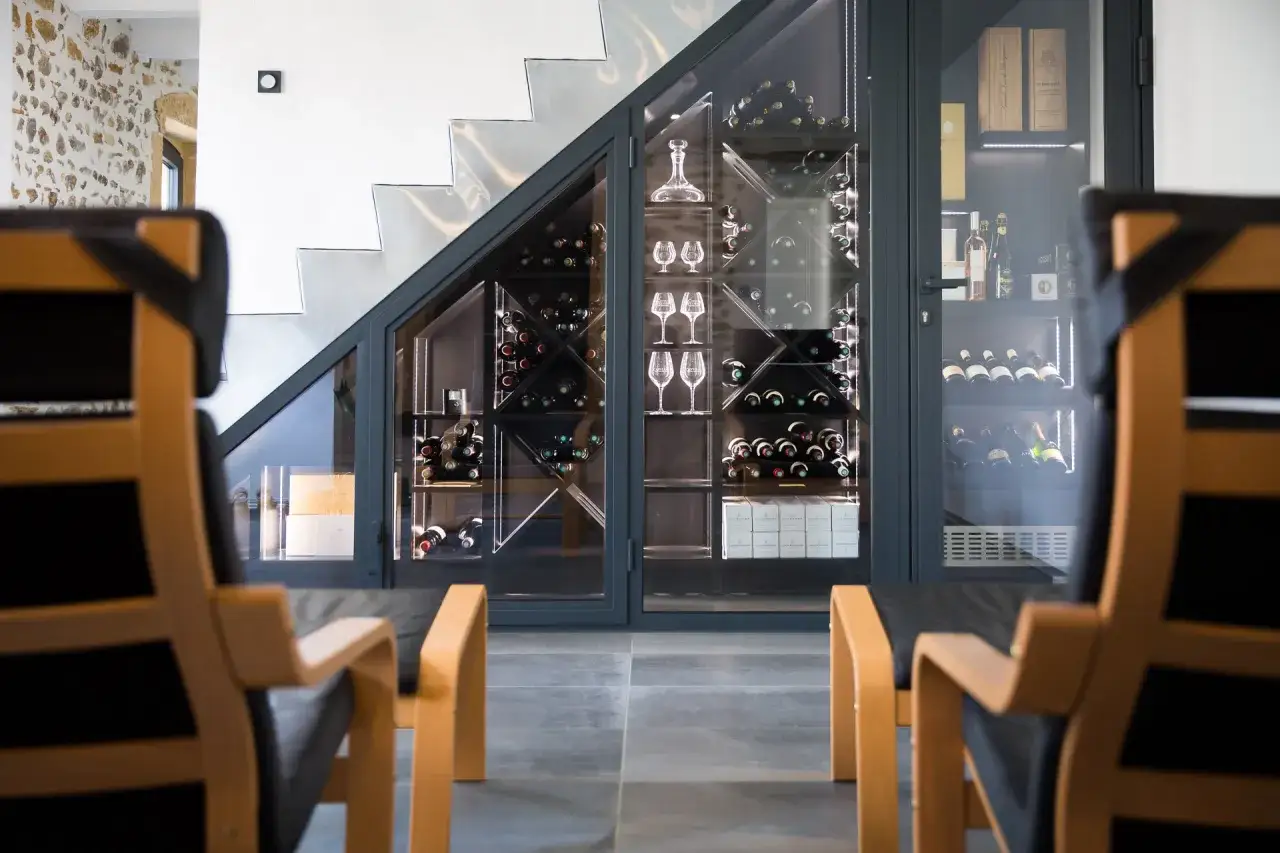 Une cave &agrave; vin moderne sous un escalier, avec des bouteilles et des verres &agrave; pied bien rang&eacute;s.