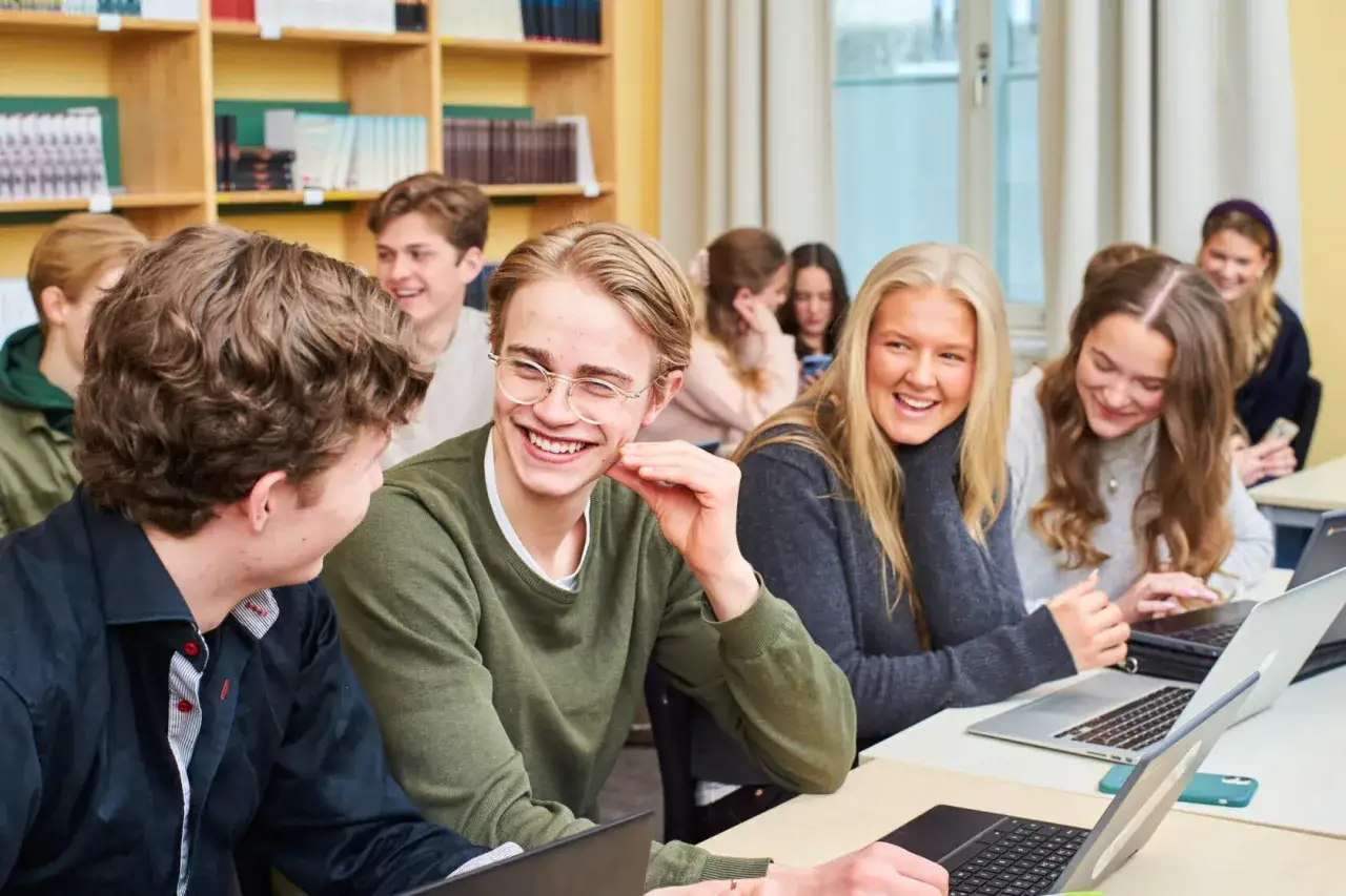 Glada elever p&aring; ett idrottsgymnasium i Stockholm skrattar tillsammans vid sina datorer i ett klassrum.