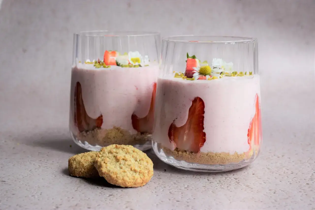 Zwei Gläser mit cremigem Erdbeer-Dessert, garniert mit frischen Erdbeeren und Gänseblümchen. Daneben liegen zwei knusprige Kekse.