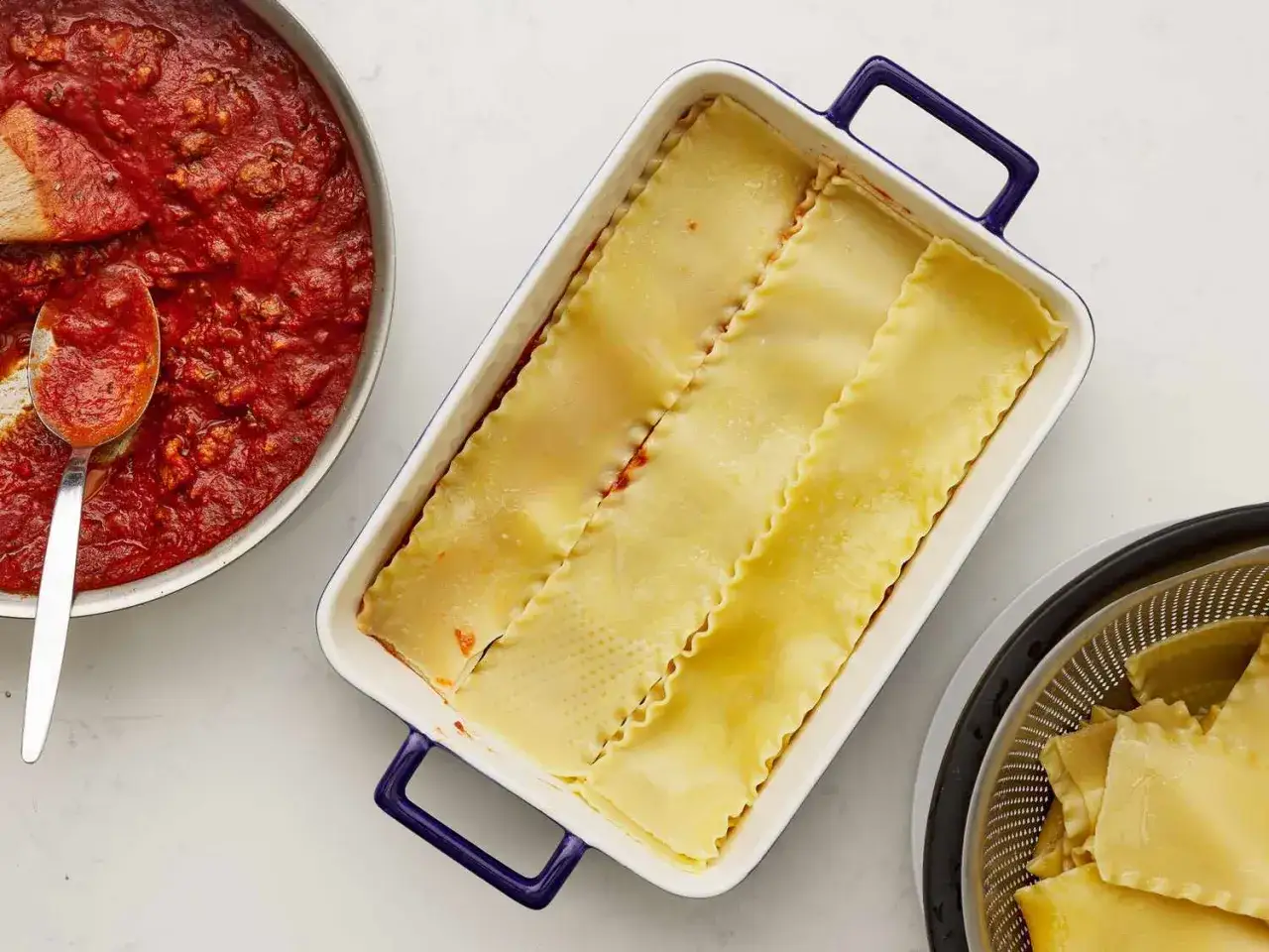 Préparation d'une lasagne maison : sauce tomate dans une poêle, pâtes dans un plat, et pâtes égouttées dans une passoire.