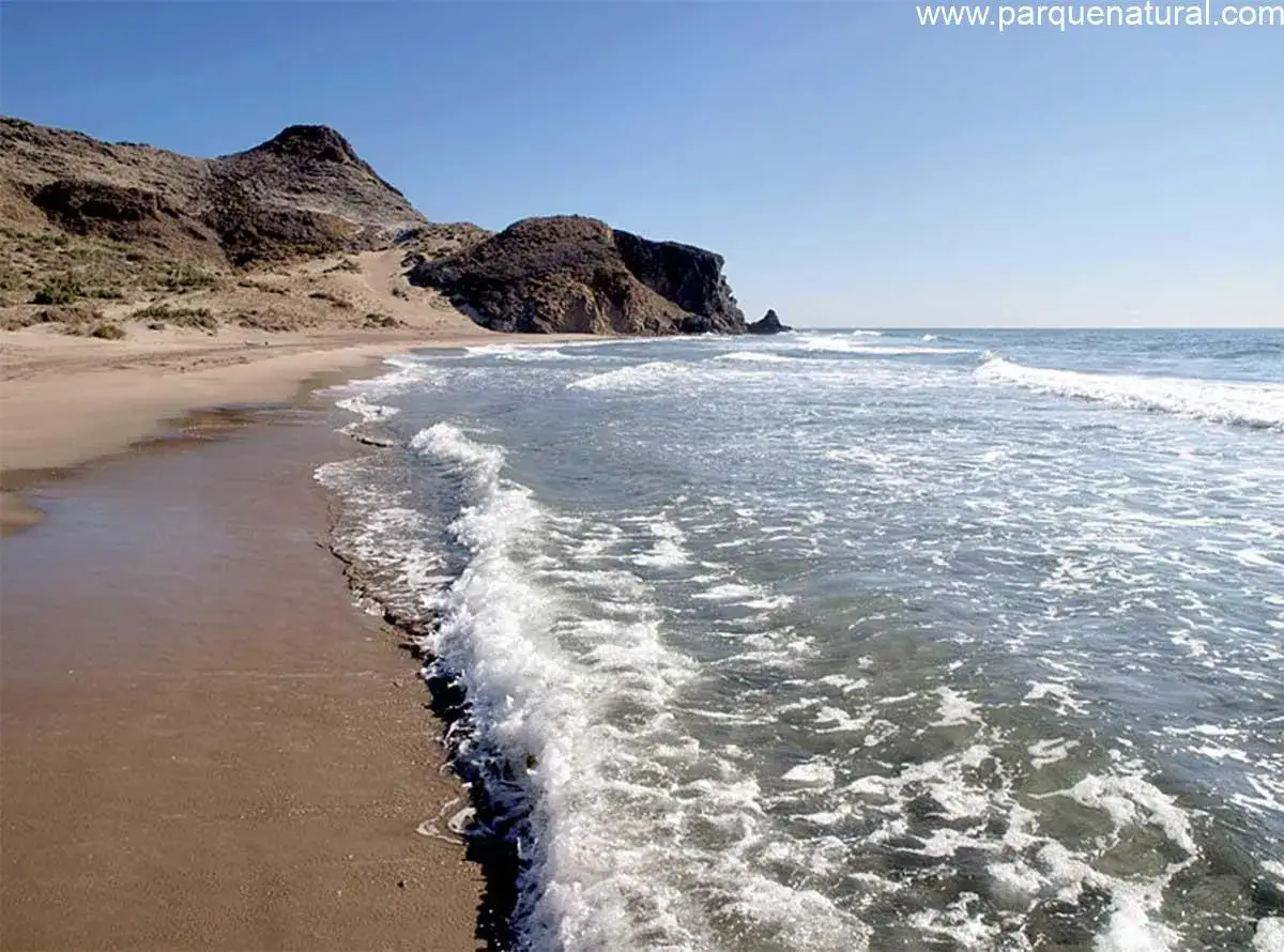 Olas rompiendo en la arena dorada de la playa del Barrronal, con formaciones rocosas al fondo bajo un cielo azul.