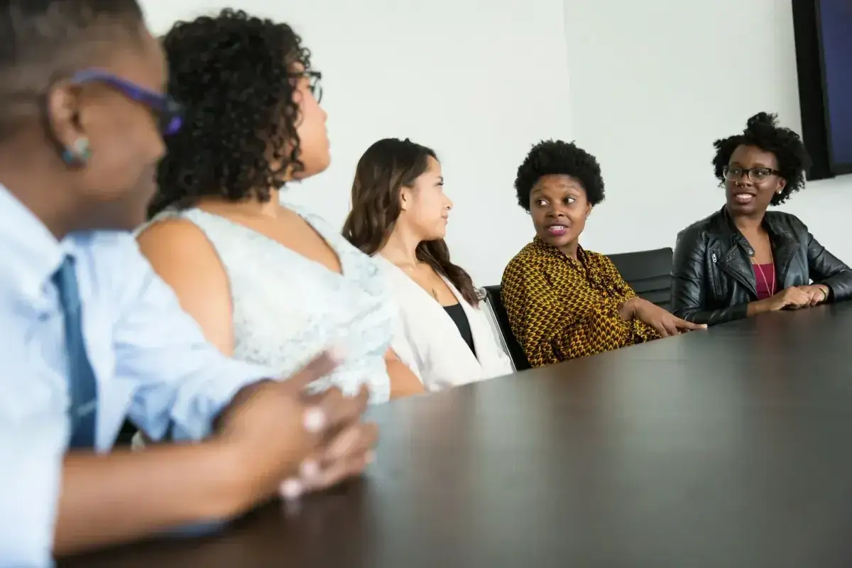 A diverse group of women in a meeting, one woman, a coach for executives, gestures while speaking to her colleagues.