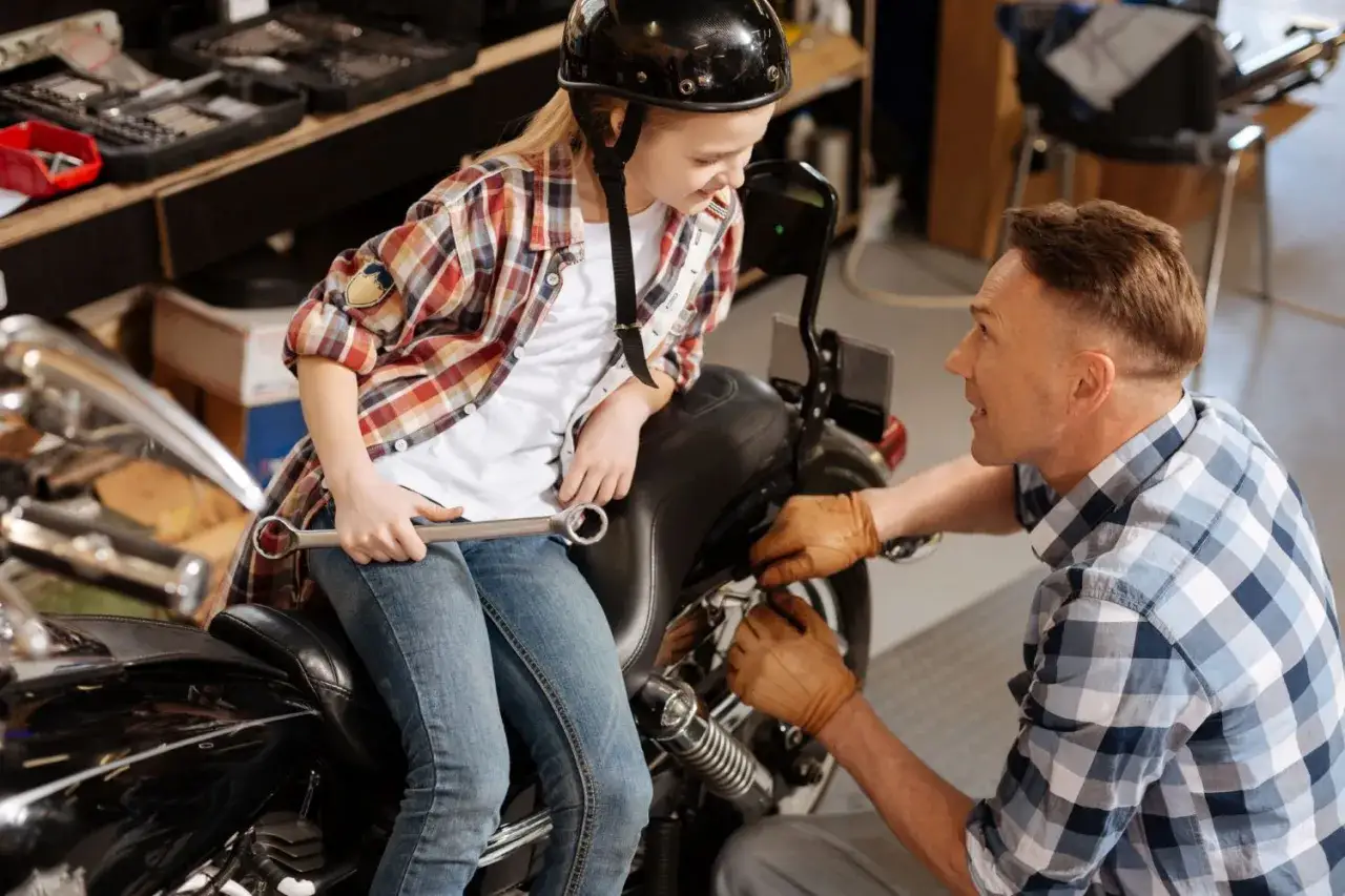 Ojciec uczy córkę, jak naprawić motocykl. Dziewczynka w kasku i kratowanej koszuli trzyma klucz, gotowa do pomocy.