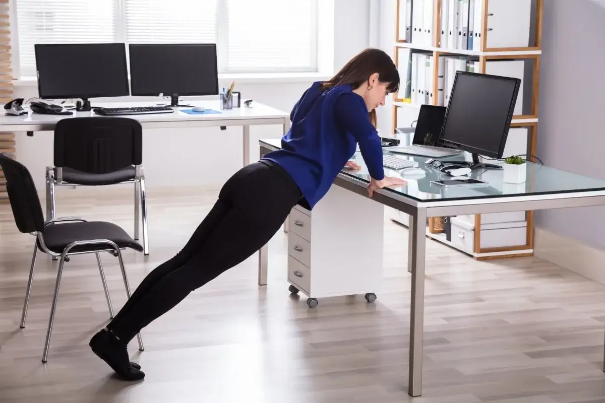 Une femme fait des pompes sur son bureau, une solution pour savoir comment faire du sport quand on a pas le temps.