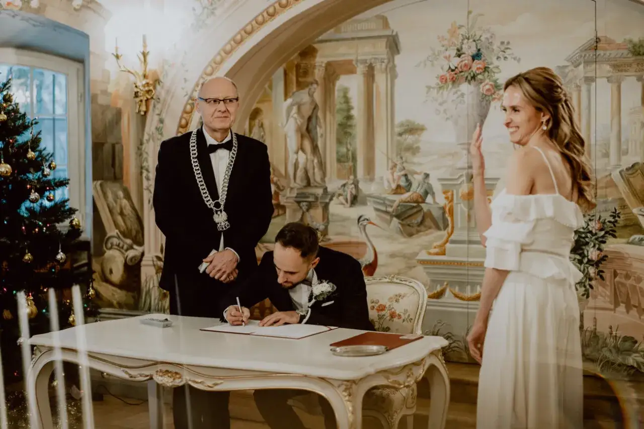 Świadek na ślubie cywilnym podpisuje dokumenty. Obok stoi urzędnik i uśmiechnięta panna młoda.