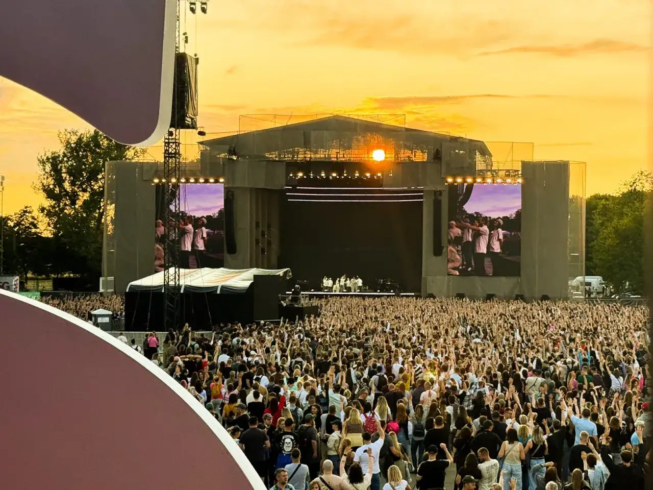 Koncert w Poznaniu pod zachodzącym słońcem. Tłum ludzi z rękami w górze, scena z ekranami.