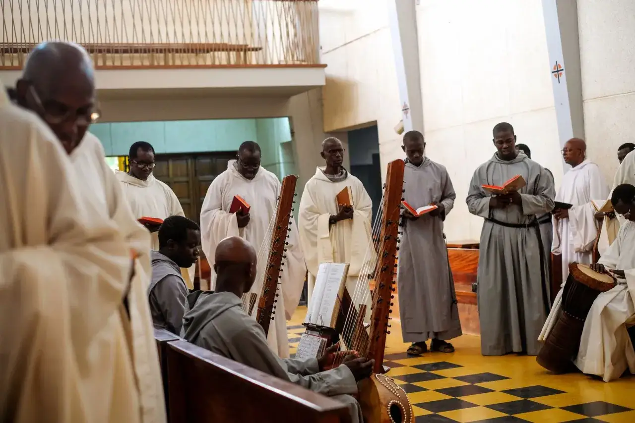 A modern day monk plays a kora harp while others in robes read from books during a service.