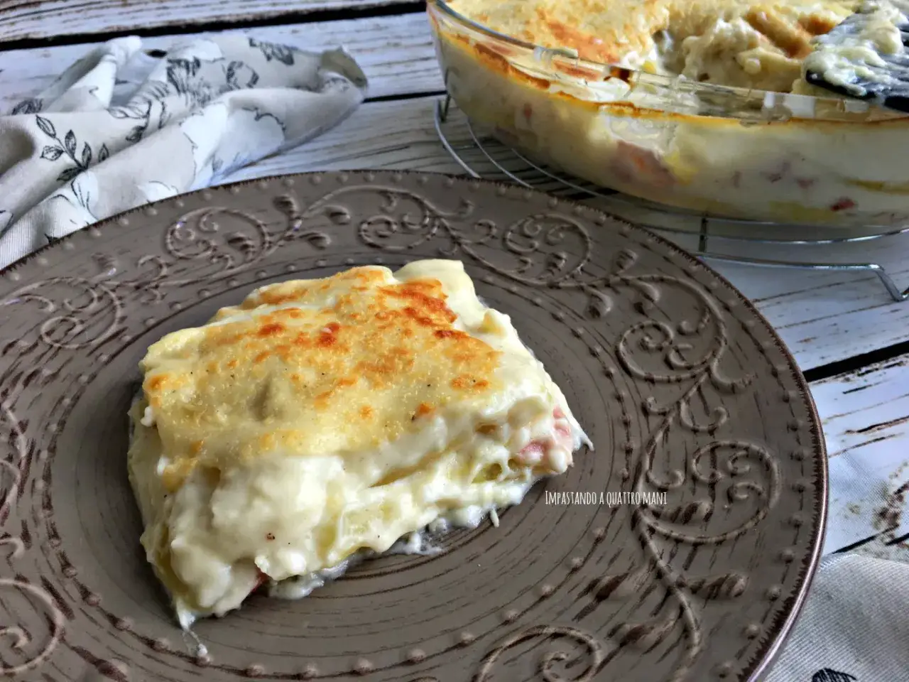 Una fetta di parmigiana di patate cremosa e dorata su un piatto decorato. In sottofondo, la teglia ancora piena.
