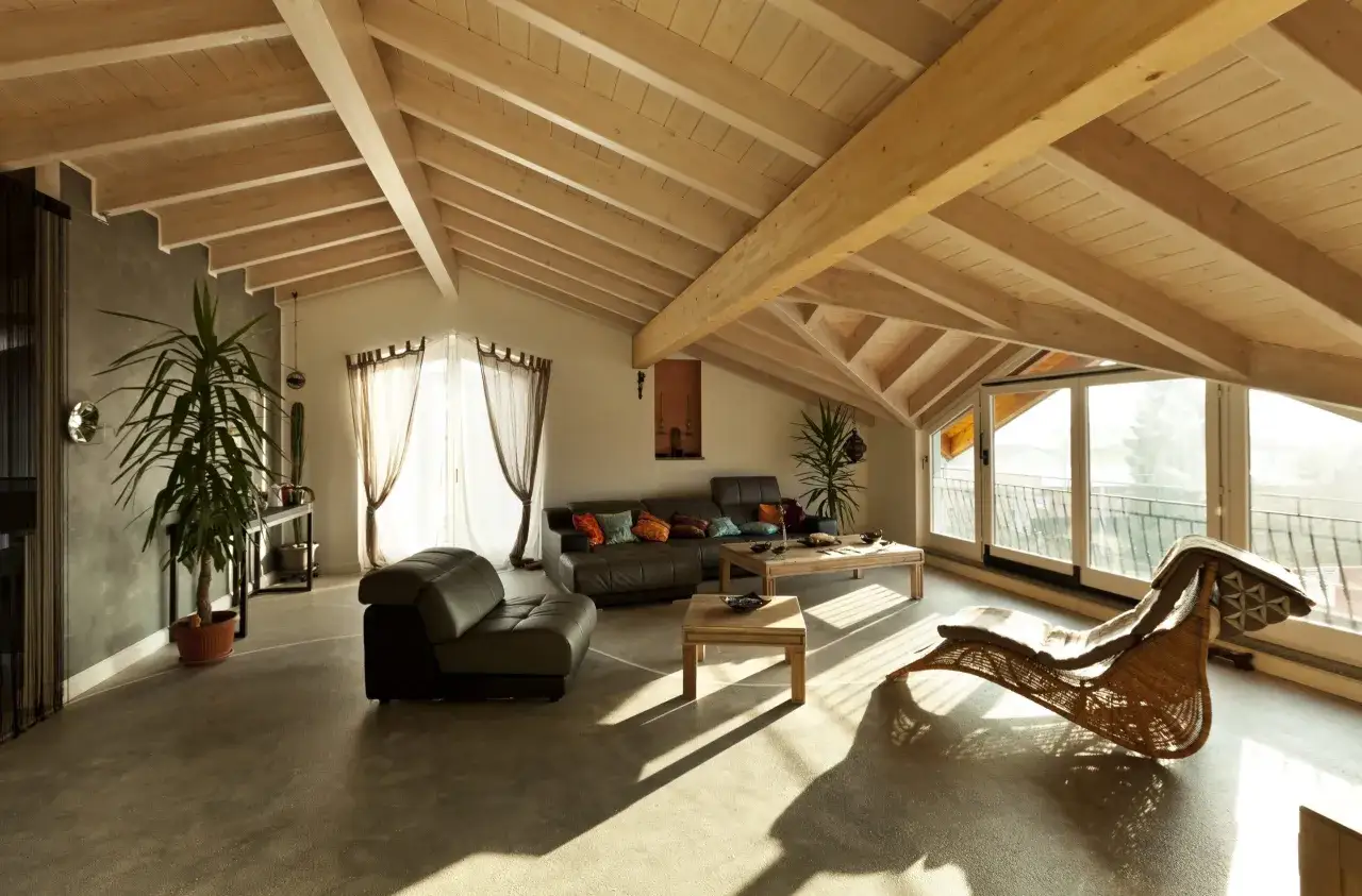 Accogliente mansarda ristrutturata con travi a vista in legno, divani in pelle, tavolini e una poltrona in vimini, illuminata da luce naturale.