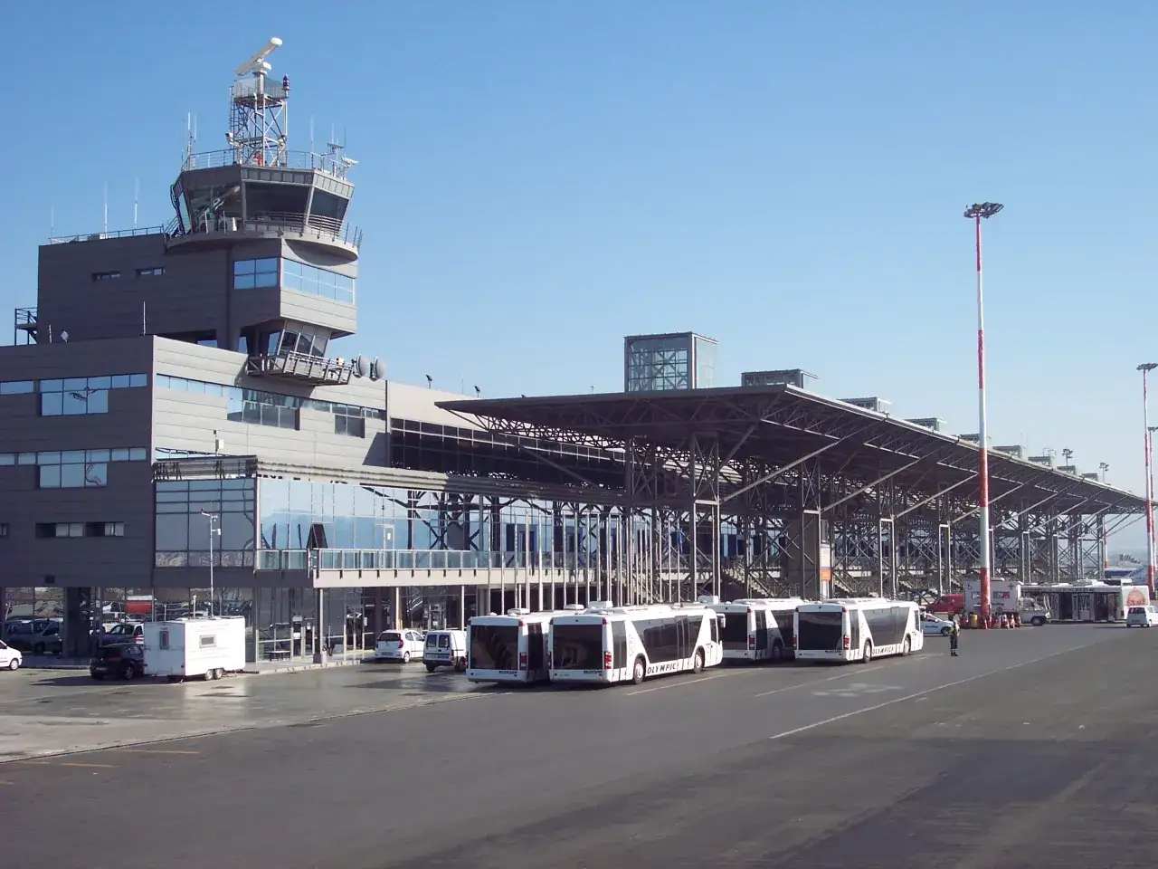 Port lotniczy Saloniki Macedonia: nowoczesny terminal z wieżą kontroli lotów, rzędem autobusów i samochodów na płycie lotniska.
