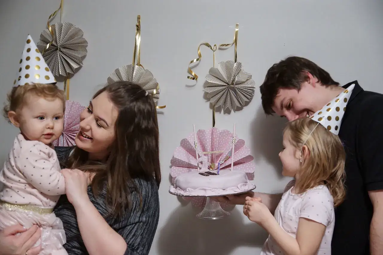 Rodzina świętuje roczek dziecka. Mama trzyma malucha, tata i starsza siostra patrzą na tort.