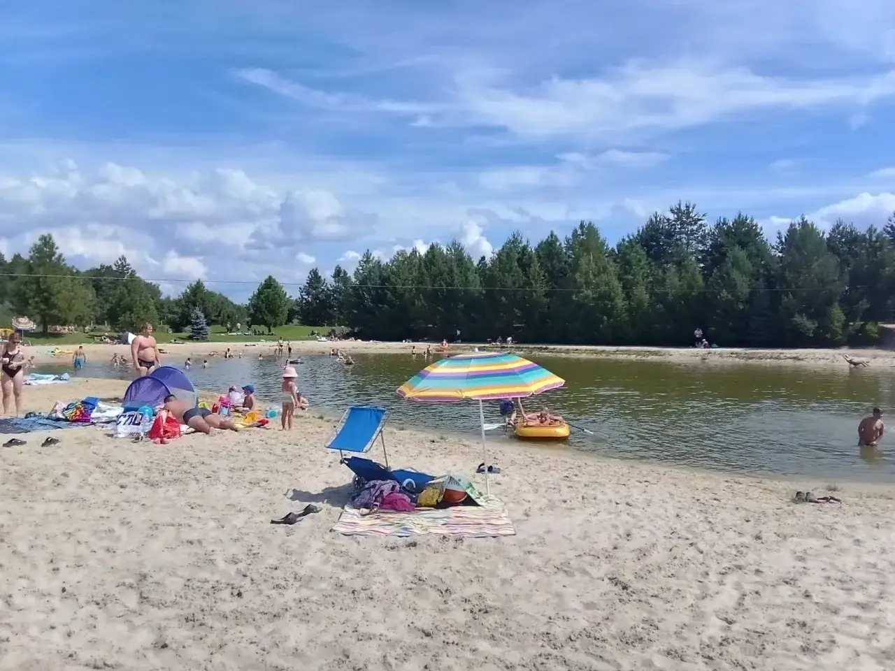 Sielska pogoda nad wodą. Ludzie odpoczywają na piaszczystej plaży, kąpią się i pływają.