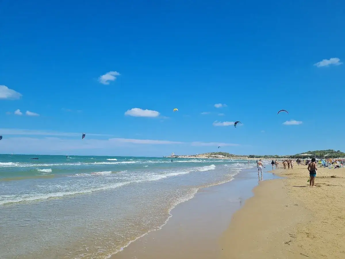 Spiaggia di Scialmarino con kitesurfer che danzano nel cielo azzurro e bagnanti che passeggiano sulla sabbia dorata.
