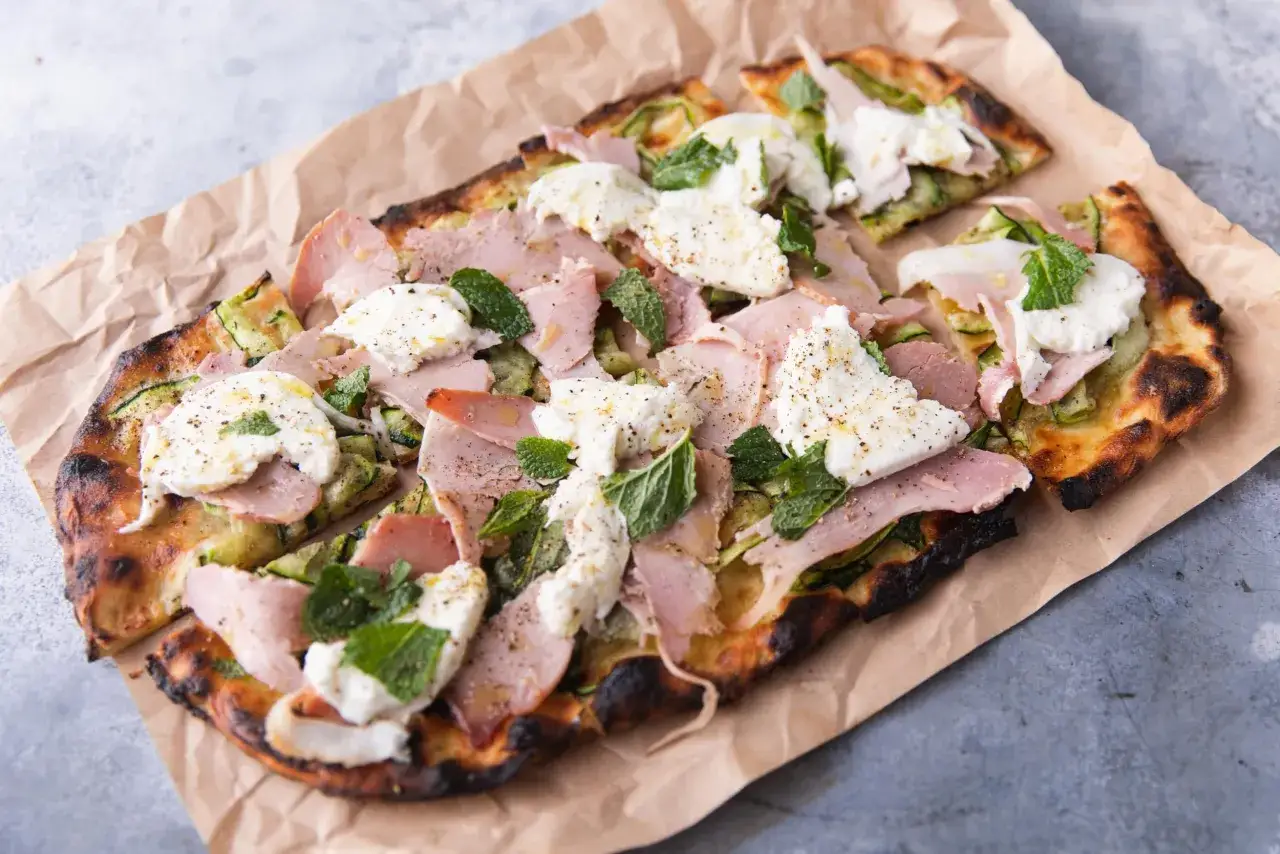 Pyszna pizza rzymska z szynką, mozzarellą, cukinią i miętą, podana na papierze.