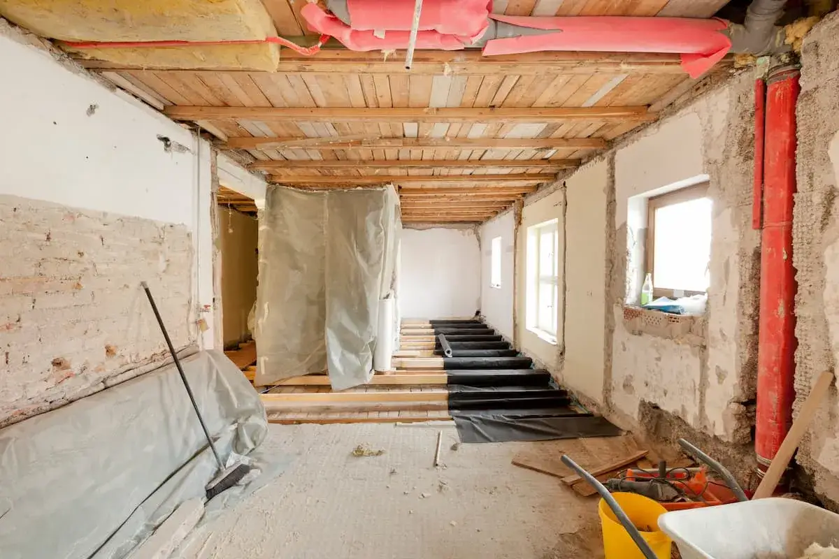 Baustelle mit freiliegenden Deckenbalken, Rohren und freigelegten Wänden. Ein Besen liegt auf abgedeckten Möbeln. Hier wird die **Sanierungspflicht** mit **Ausnahmen** umgesetzt.