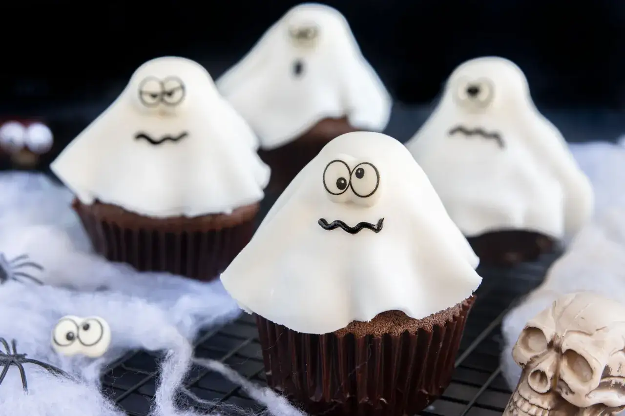 Vier schaurig-schöne gespenster muffins mit weißen Geisterhauben und lustigen Gesichtern, dekoriert mit Spinnweben und einem Totenkopf.