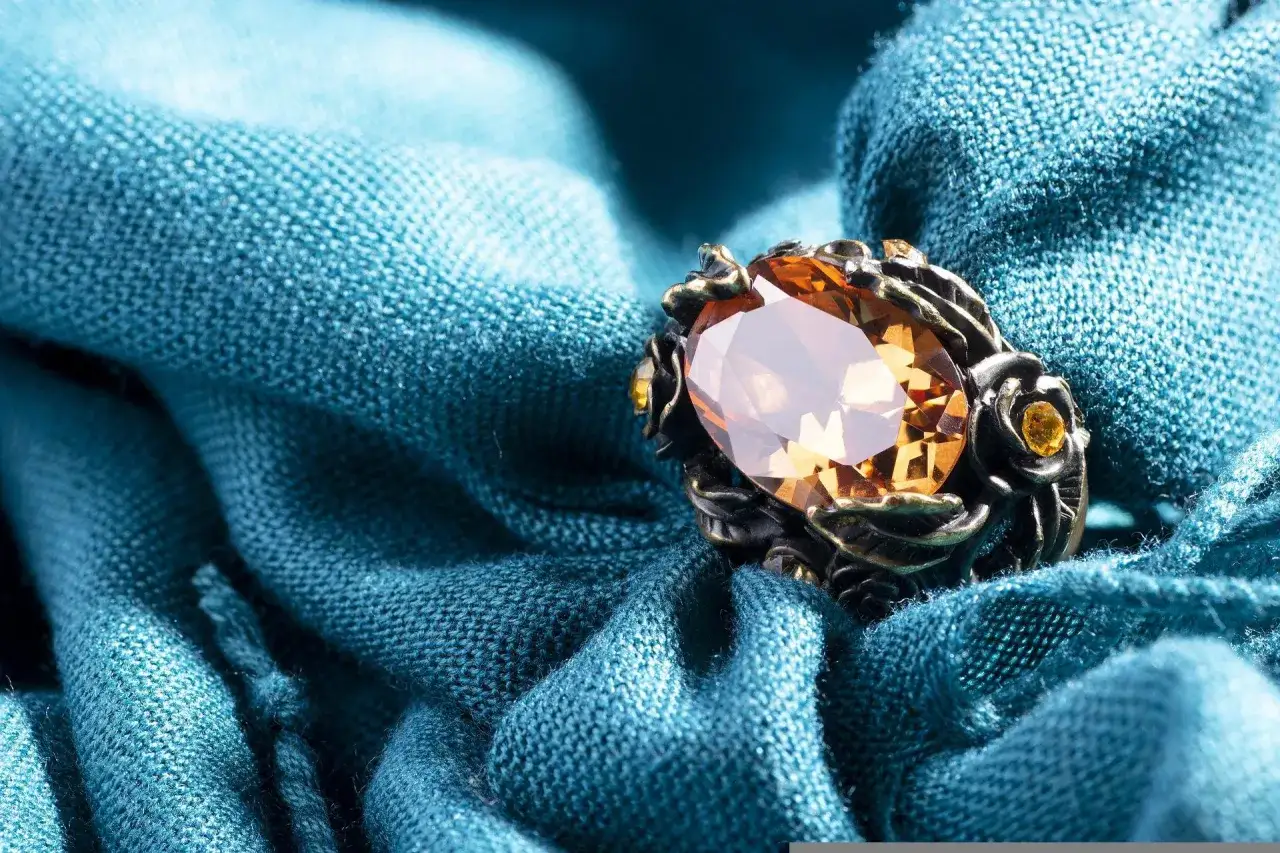 Ein Ring mit einem großen, ovalen, bernsteinfarbenen Stein und kleineren gelben Steinen, eingebettet in ein dunkles Metallgehäuse, liegt auf einem türkisfarbenen Stoff.