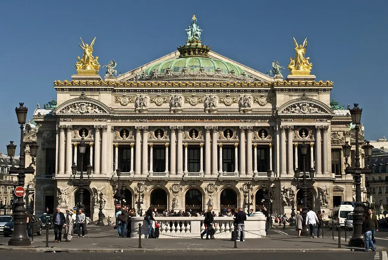 La **place de l'Op&eacute;ra** &agrave; Paris, avec son imposant b&acirc;timent orn&eacute; de statues dor&eacute;es et d'une coupole verte, sous un ciel bleu &eacute;clatant.